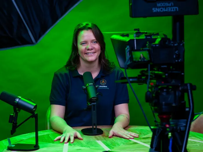 A host seated at the studio table against the green screen, ready for keying